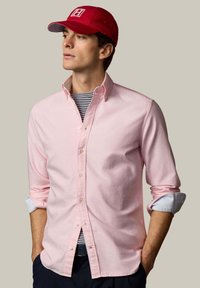 Muž v červenej šiltovke, svetloružovej košeli s gombíkmi s vyhrnutými rukávmi, pruhovanom tričku pod ňou a tmavých nohaviciach, ruky vreckách, pozerá sa doľava.