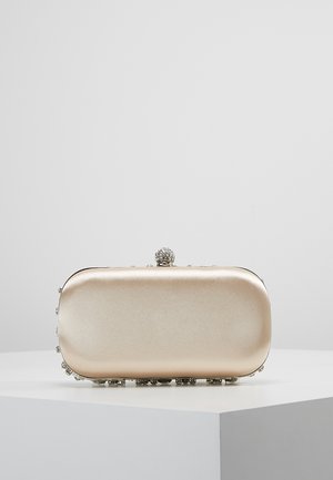 Kleine rechthoekige gouden clutch met afgeronde hoeken, bovenop een sierlijke sluiting met juwelen, rustend op een wit voetstuk tegen een effen achtergrond.