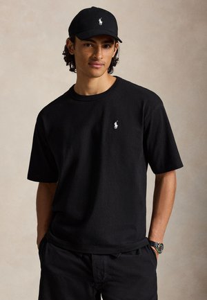 Noormees keskmise nahatooniga, kandes musta Polo Ralph Laureni nokamütsi, musta t-särki valge logoga, musti pükse ja käekella, käed taskutes.