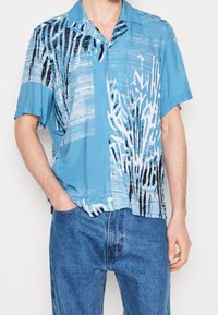 Koszula z krótkim rękawem wykonana z jasnoniebieskiej tkaniny z abstrakcyjnymi czarno-białymi wzorami, o luźnym kroju, z zapięciem na guziki i kołnierzykiem.