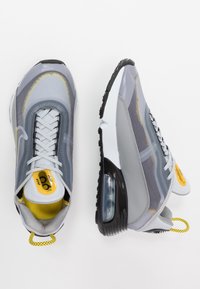 Grå atletiska sneakers med mesh- och syntetiska material, med en synlig luftenhet, gula accenter och svarta gummisulor.