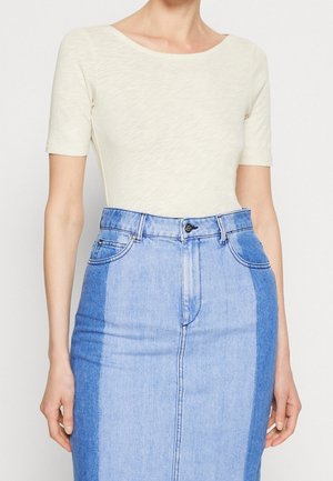 Frau trägt ein cremefarbenes Kurzarmtop, das in einen hochtaillierten blauen Denimrock mit dunklen Seitenteilen und vorderen Taschen gesteckt ist.