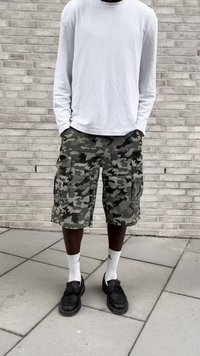 Person wearing white long-sleeve shirt, camo cargo shorts, white Adidas socks, and black tassel loafers standing on tiled ground near a brick wall.