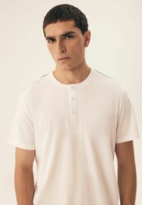 White short-sleeve henley shirt featuring a round neckline and three buttons at the front. Smooth fabric with subtle side accents.