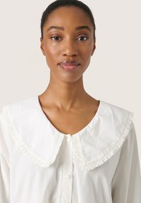 Blusa di cotone bianco con colletto rotondo arricciato, chiusura con bottoni e texture liscia. Un design classico con un aspetto pulito e minimalista.