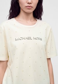 Femme portant un t-shirt de couleur crème avec de petites clous dorés et le logo "MICHAEL KORS" imprimé sur la poitrine.