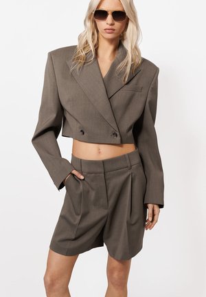 Femeie purtând un sacou gri supradimensionat croptat, pantaloni scurți cu talie înaltă asortați și ochelari de soare, pozând cu o mână în buzunar pe un fundal alb.