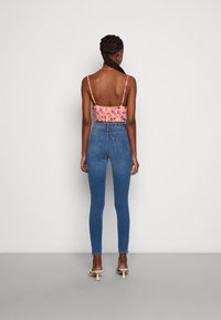 Os jeans azuis skinny apresentam uma cintura alta, dois bolsos traseiros e uma textura de denim suave. O modelo usa uma blusa com padrão floral e alças finas.