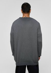 Pull en tricot gris avec une coupe ample, présentant une texture côtelée, un col rond et des fentes latérales. Fini avec des épaules tombantes.