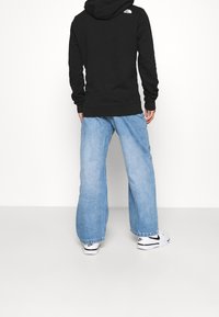 Felpa nera con cappuccio, tasca a marsupio e logo sul retro, abbinata a jeans a gamba larga azzurri e sneakers bianche con dettagli neri.