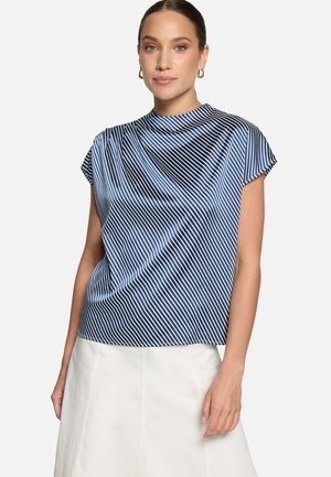 Kobieta ubrana w niebiesko-biały, diagonalnie paskowany, bezrękawnik z wysokim kołnierzem oraz białą spódnicę, stojąca na jednolitym tle.