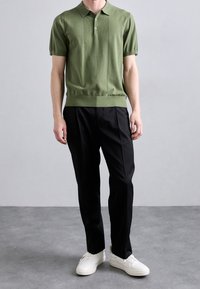 Hombre vestido con polo de punto de manga corta en verde oliva, pantalones negros a medida y zapatillas blancas, de pie sobre un suelo gris contra una pared lisa.