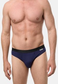 Męski tors w granatowych majtkach z czarnym paskiem w talii i małą metką po prawej stronie, stojący na jasnym, jednolitym tle.