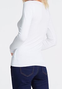 Eine Frau trägt ein enges weißes Langarmoberteil und dunkelblaue Jeans, von hinten gezeigt, mit sichtbarem blonden Haar.