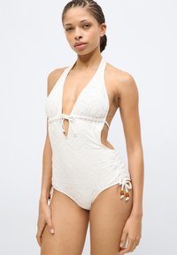 Λευκό ολόσωμο μαγιό με υφή από σχέδιο, βαθύ V λαιμόκοψη και ανοίγματα στα πλάγια. Διαθέτει ρυθμιζόμενους δεσίματα και διακοσμητικά χάντρες στα πλάγια.