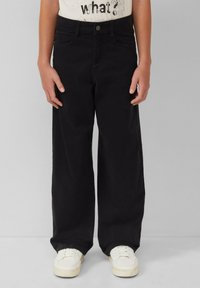 Persona que lleva pantalones de pierna ancha negros de talle alto, zapatillas blancas y una camiseta blanca con el texto "¿qué?", de pie contra un fondo liso.