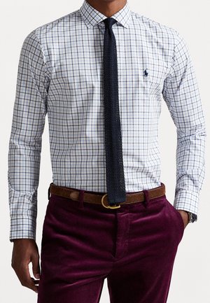 Man draagt een wit/blauw geruit overhemd, een donker marineblauwe gebreide stropdas, een bruine riem en donkerpaarse corduroy broek met hand in de zak.