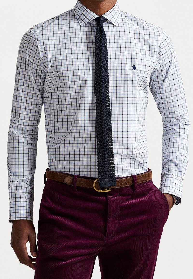 Homme portant une chemise à carreaux blanche et bleue, une cravate tricotée bleu marine foncé, une ceinture marron et un pantalon en velours côtelé violet foncé avec la main dans la poche.