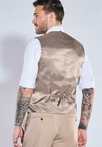 Taubengraue Satweste mit glatter Textur, verstellbarer Schnalle auf der Rückseite, getragen über einem weißen Langarmhemd, kombiniert mit beigen Hosen.