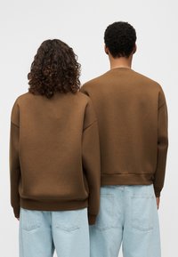 Καφέ φούτερ με χαμηλωμένους ώμους, ριμπ μανσέτες και ζώνες μέσης, συνδυασμένα με ανοιχτόχρωμα μπλε τζιν, εμφανιζόμενα από πίσω.