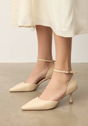 Beige spitze High Heels mit Knöchelriemen, getragen zu einem leichten, fließenden Rock, stehend auf einem glatten, grauen Boden.