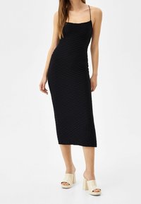 Schwarzes Bodycon-Kleid mit geripptem Textur, Spaghetti-Trägern und Midi-Länge. Kombiniert mit nude-farbenen High-Heels.