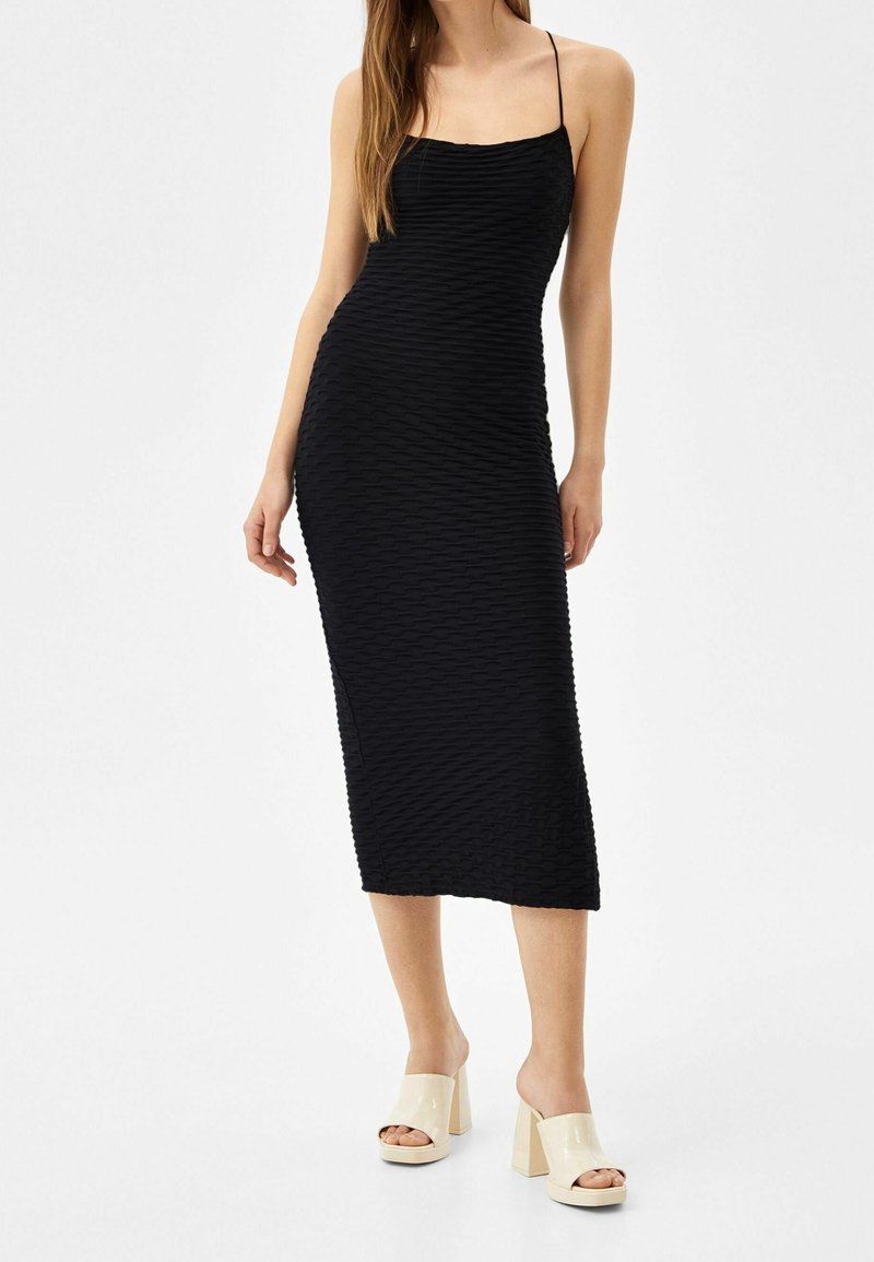 Schwarzes Bodycon-Kleid mit geripptem Textur, Spaghetti-Trägern und Midi-Länge. Kombiniert mit nude-farbenen High-Heels.