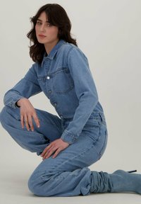 Lichtblauw denim jumpsuit met een kraag, zakken aan de voorkant en knoopsluitingen. Losse pasvorm met taps toelopende benen.