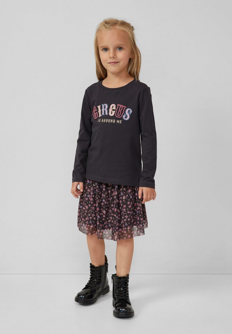 Een jong meisje draagt een zwarte lange mouwen shirt met de tekst "CIRCUS is around me", een bloemenrok en zwarte glanzende veterschoenen terwijl ze op een grijze vloer staat.