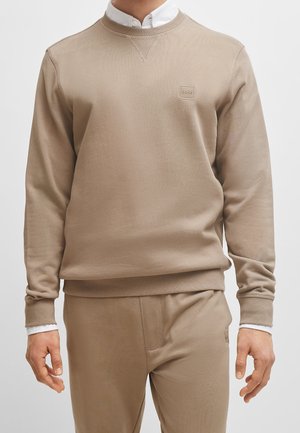 Mężczyzna w beżowej bluzie z okrągłym dekoltem i pasujących spodniach, nałożonych na białą koszulę z kołnierzykiem, stojący na jednolitym tle.