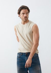Beige sleeveless t-shirt made of soft fabric, paired with dark blue denim jeans. The styling features a relaxed fit and subtle stitching details.