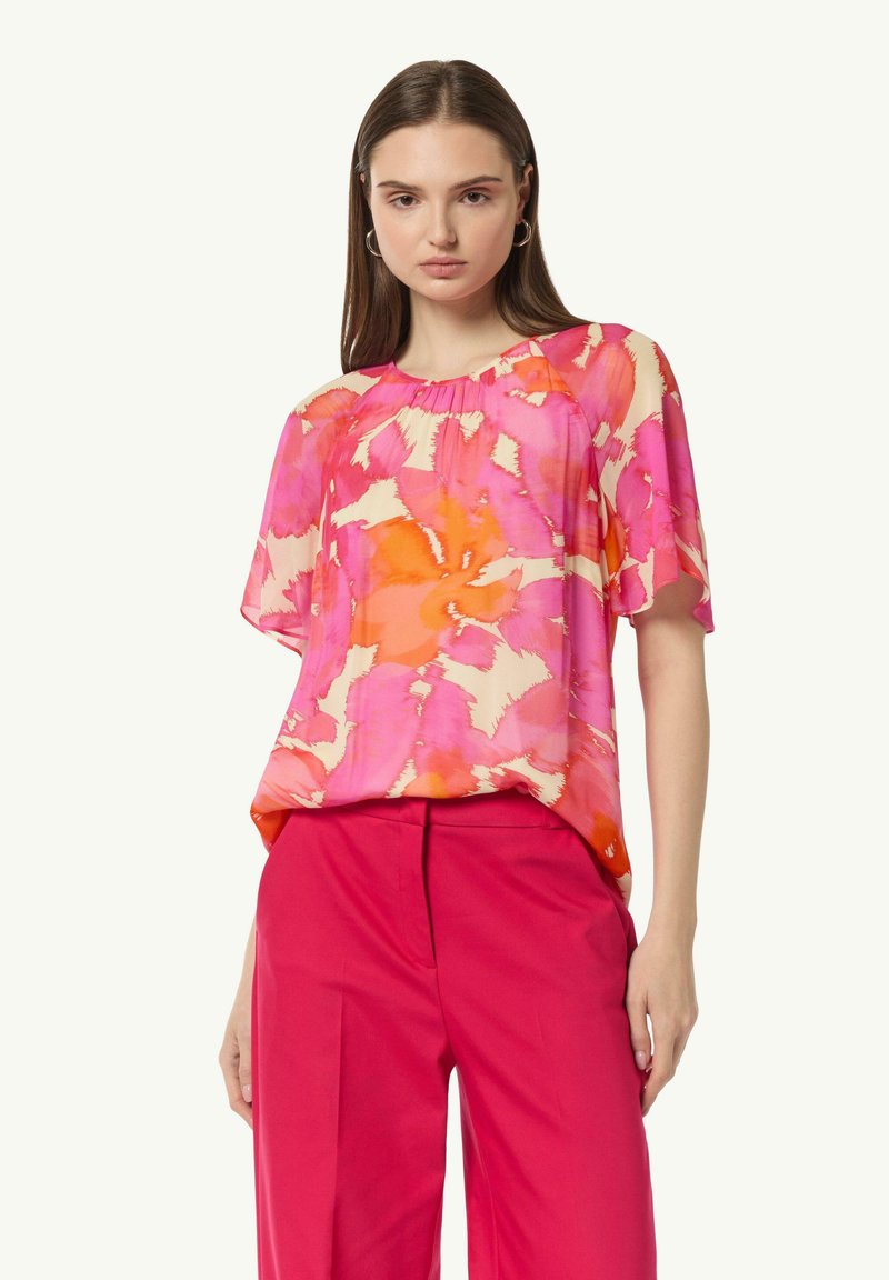 Bluzka w kwiatowy wzór w odcieniach różu i pomarańczu, o luźnym kroju, z krótkimi rękawami i marszczonym dekoltem. Połączona z różowymi spodniami.