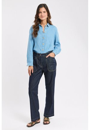 Femme aux longs cheveux portant une chemise bleu clair boutonnée, un jean ample à larges jambes avec de grandes poches, et des sandales foncées, debout devant un fond blanc.