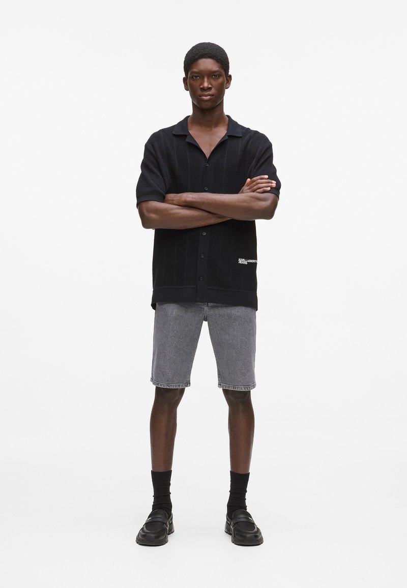 Schwarzes kurzärmeliges Hemd mit vertikaler Rippenstruktur und Knöpfen, kombiniert mit grauen Denim-Shorts, schwarzen Socken und schwarzen Schuhen.