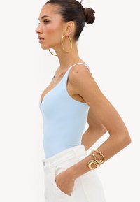 Femme aux cheveux foncés attachés en chignon porte un top sans manches bleu clair, de grandes boucles d’oreilles dorées, un pantalon blanc et un bracelet épais en or à son bras droit.