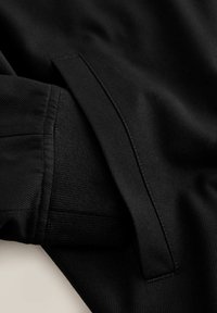 Fekete ruhaujj közeli kép ribbedezős mandzsettával és varrott zsebbel, texturált anyagon.