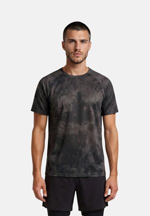 Jonge man met kort haar die een zwart-grijs tie-dye sportshirt en zwarte korte broek draagt, staand tegen een lichte achtergrond.