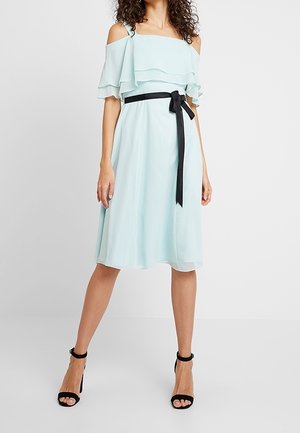 Kvinde iført en lyseblå off-shoulder midi-kjole med flæsedetalje og sort taljebånd, stylet med sorte højhælede sandaler.