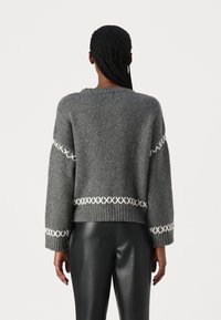 Grauer gestrickter Pullover mit lockerer Passform, weiten Ärmeln und weißen Kreuzstichakzenten an den Bündchen und dem Saum. Rippstrickbund und Struktur.