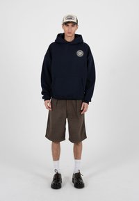 Tânăr îmbrăcat într-un hanorac bleumarin cu logo, pantaloni scurți maro până la genunchi, șosete albe, pantofi negri și o șapcă bej, sprijinit de un fundal simplu.