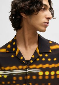 Short-sleeved shirt featuring a black fabric with yellow and white abstract patterns, a collar, and a smooth texture.