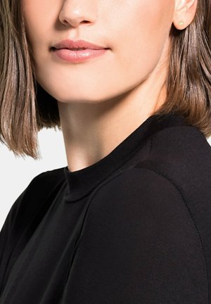 Czarny top z wysokim kołnierzem, wykonany z miękkiego materiału, o gładkiej fakturze i detalach na szwach ramion. W uszach noszone są złote kolczyki wkręty.