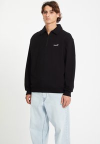 Sweat à fermeture éclair noir avec un col, arborant un petit logo blanc sur la poitrine. Associé à un jean baggy bleu clair.