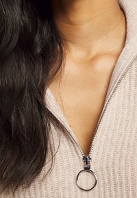 Beiger Strickpullover mit einem silbernen Reißverschluss und einem runden Zipper-Anhänger. Nahaufnahme des Halsausschnitts und Schulterbereichs, die die Stoffstruktur hervorhebt.