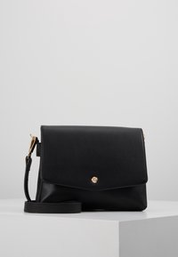 Černá kožená kabelka přes rameno s klopou a zlatým patentním uzávěrem, umístěná na bílé podstavě proti jednolité šedé kulise.