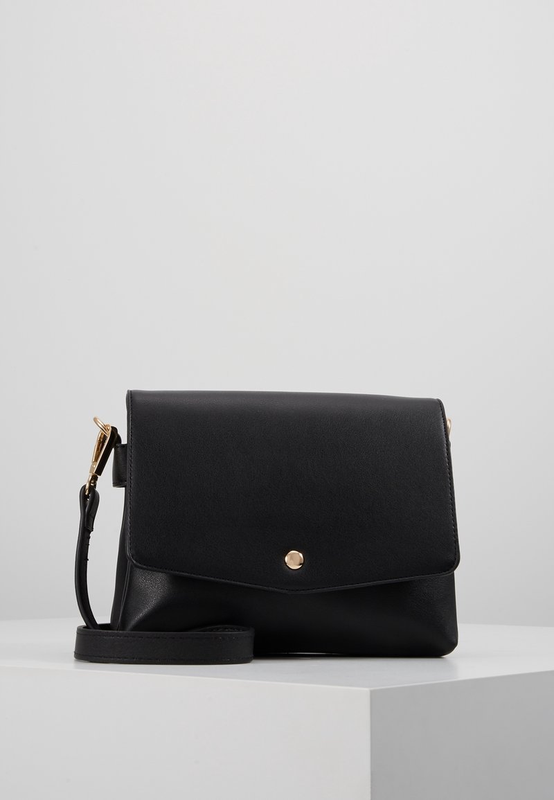 Černá kožená kabelka přes rameno s klopou a zlatým patentním uzávěrem, umístěná na bílé podstavě proti jednolité šedé kulise.