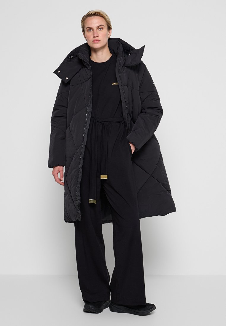 Schwarze Steppjacke mit einer gepolmerten Optik, ausgestattet mit einer Kapuze und Reißverschlüssen. Getragen über einem schwarzen Outfit mit Kordelhose und flachem Schuhwerk.