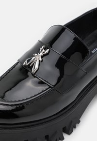 Mocassin en cuir verni noir brillant avec semelle épaisse à crampons et ornement métallique argenté en forme d’insecte sur le dessus.