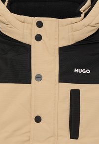 Tan in črna podložena jakna z visokim ovratnikom, opremljena s črnimi pritiskači in žepom na prsih, blagovno znamko "HUGO" v belih črkah.