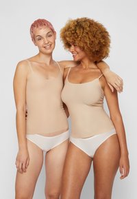 Deux femmes portent des débardeurs ajustés couleur beige clair avec des fines bretelles, associés à des sous-vêtements blancs. Le tissu semble lisse et extensible.